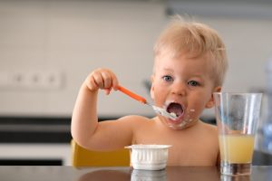 Adorable one year old baby boy eating yoghurt with spoon. Dirty messy face of toddler child.